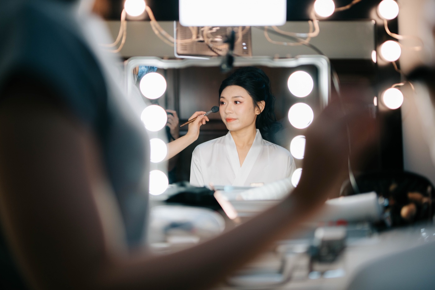 Make-up artist brushing on a bride’s make-up at a lit vanity mirror